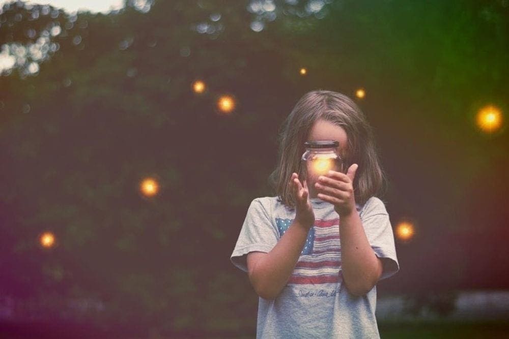 A young firl with an American flag shirt on holds a mason jar with a lightnight bug in it.