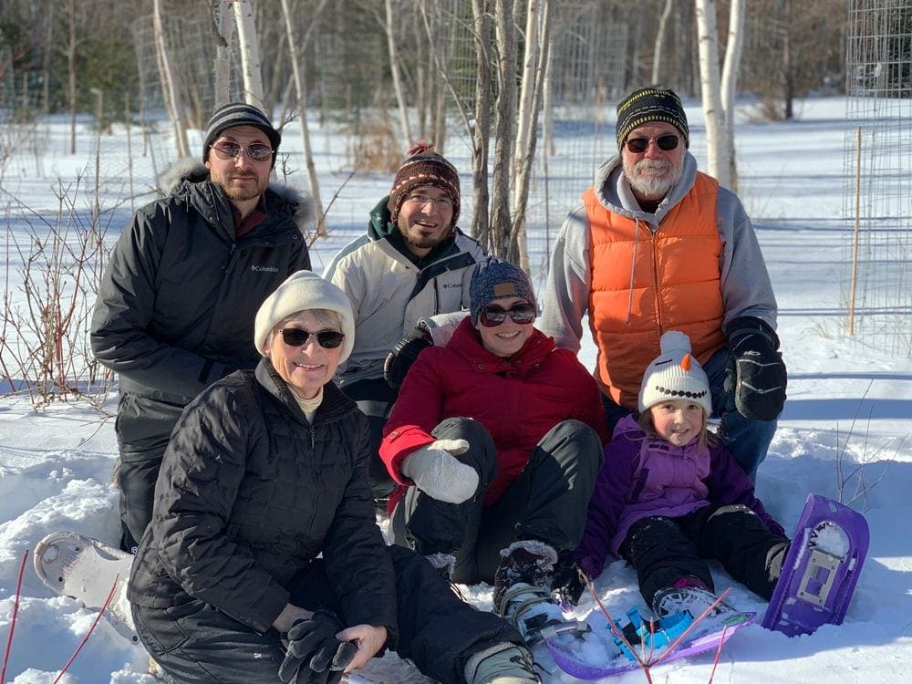 Five adults and one young child, all in snow gear and snowshoes, sit in the snow with big smiles.