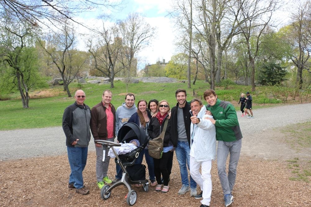Nine adults smile proudly and surround a baby carriage in a NYC park.