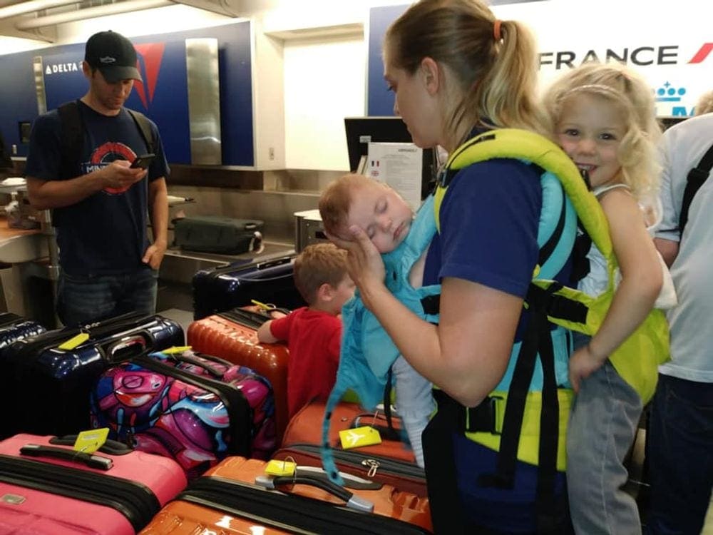 A mom wears an infant on her chest and a toddler on her back as she surveys their luggage and dad looks at his phone.