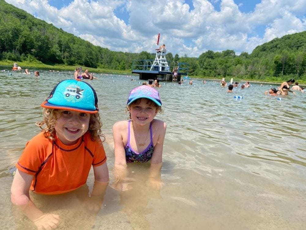 Two kids lean in close as they are laying in the water at Belleayre Beach. A lifeguard stand and other swimmers can be seen in the background. Belleayre Beach is one of the best family-friendly beaches in the Catskills!