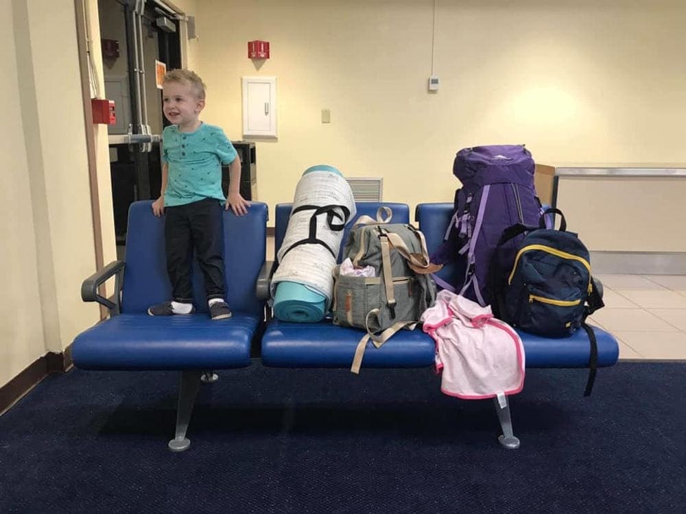 A toddler boy stands on a blue waiting room chair in the airport, while a mound of luggage filles the remaining two seats.