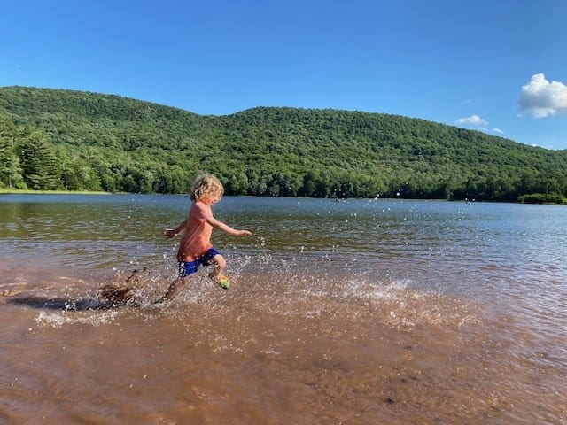 A young child wearing an orange shirt runs through shallow waters at Colgate Lake creating huge splashes around his legs.