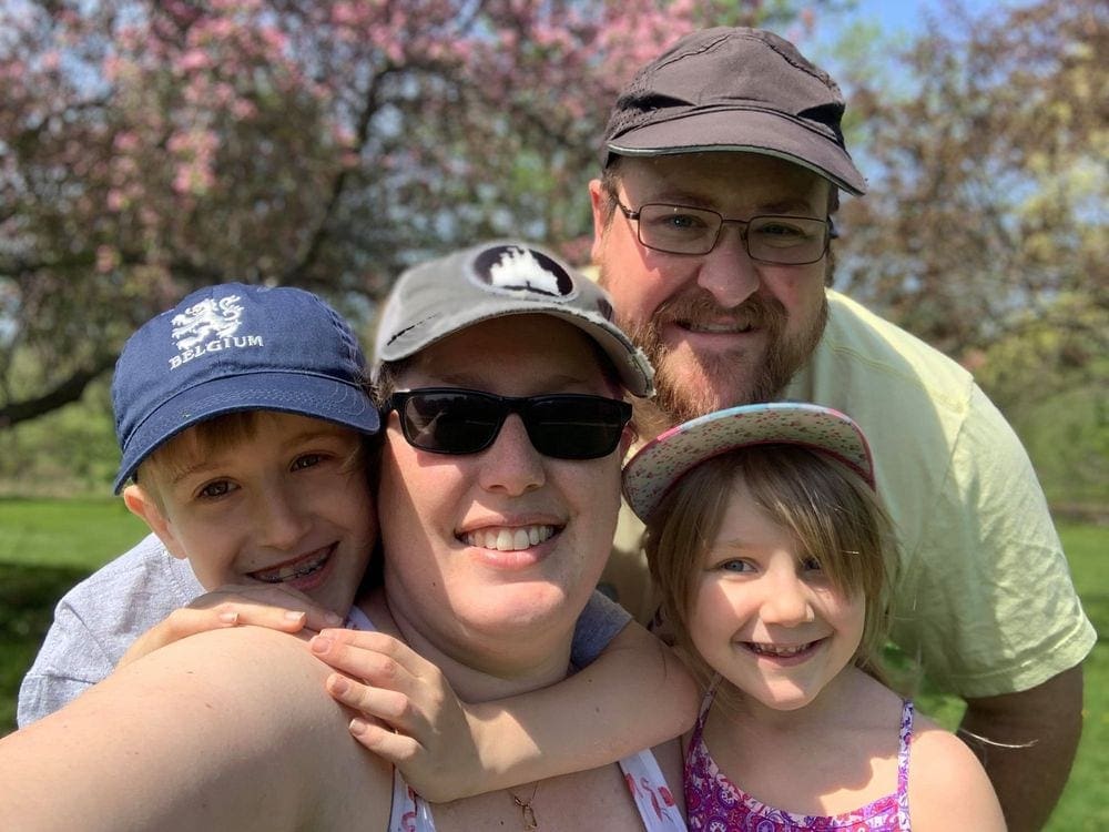 A family of four huddles together as the mom takes a selfie of them, with cherry blossoms in the background.