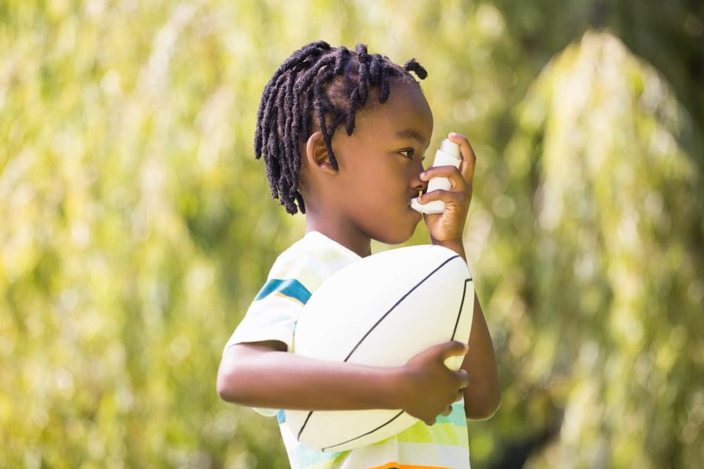 A young black child self-administers an inhaler while holding a football in his other hand.