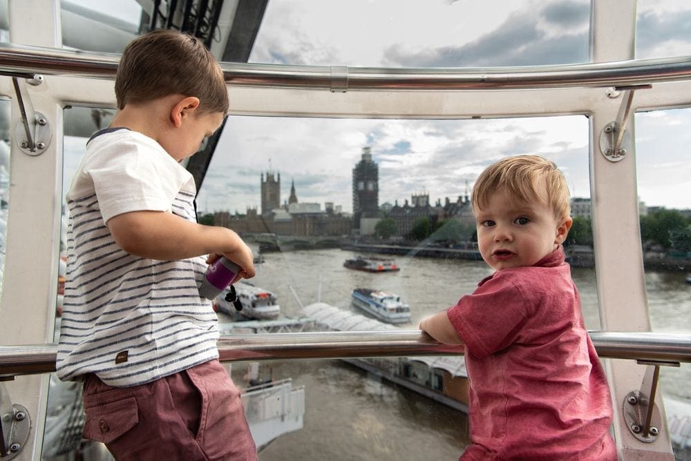 A young boy looks outside of their pod on the London Eye, while his younger brother looks back at the camera. Outside of the pod, the Thames River and Big Ben are shown in the distance.