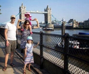 A happy family of four stands in front of the London Bridge, the youngest daughter rides on the shoulders of her mom.
