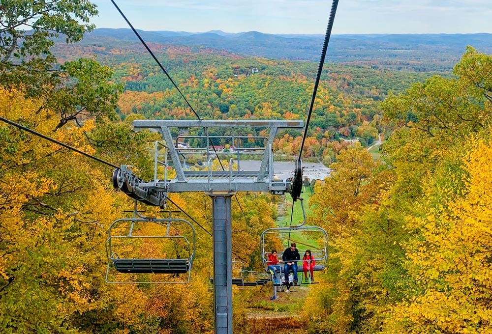A ski chair hold an adult and two children amongst sweeping views of the golden fall foliage in the Berkshires of Massachusettes, one of the best places for fall foliage in New England.