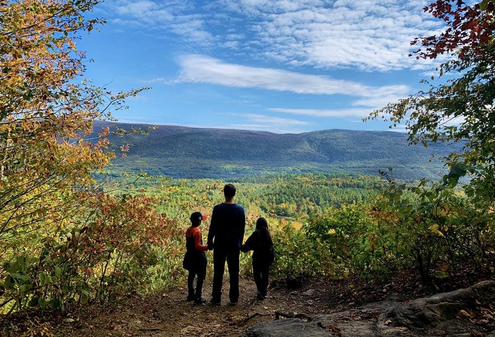 An adult holds the hands of two small chidlren wile standing on the edge of a cliff overlooking an incredible fall scene on a brilliantly sunny day.