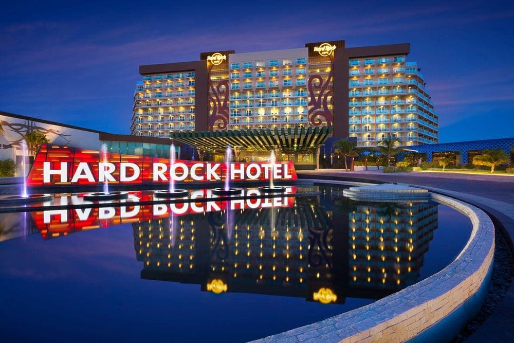 A view of the front of the Hard Rock Hotel Cancun, including a water feature and a sign of the name in white with a red background.