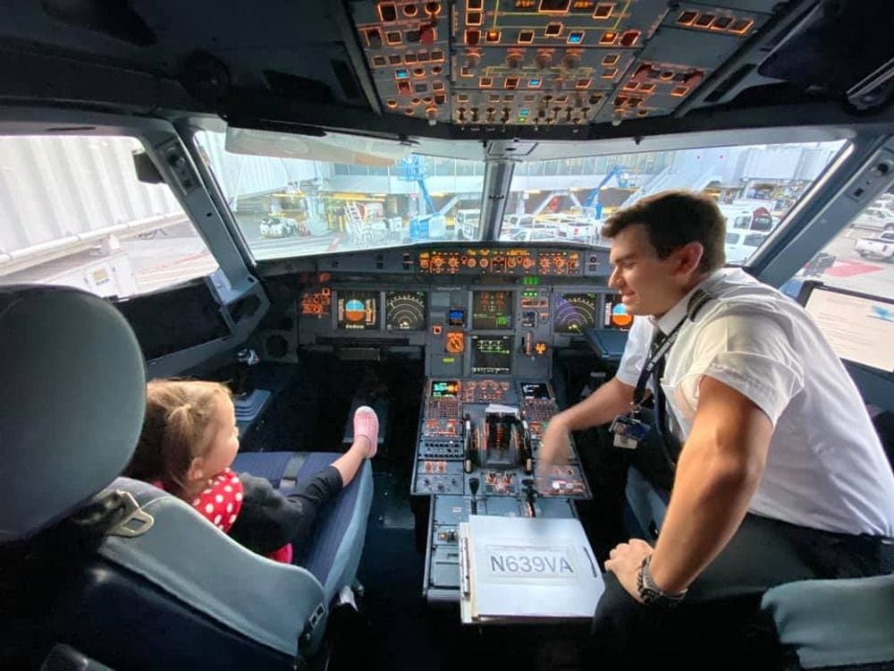 A pilot leans over to speak with an infant girl who's sitting in the cockpit.