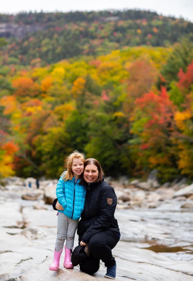 A mom and her young daughter stand together amongst the fall foliage of New Hampshire.