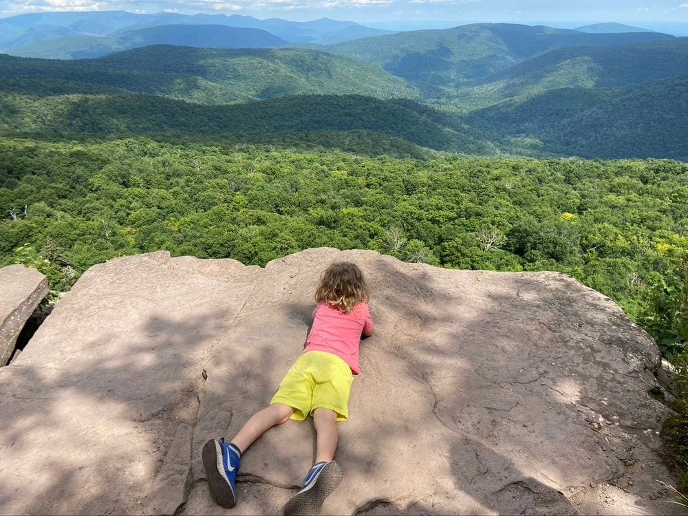 A young child lays on his stomach on a rock ledge while looking out at an expansive view of the Catskill Mountains at the top of the Giant Ledge Trail, one of the most kid-friendly hikes in the Hudson Valley.