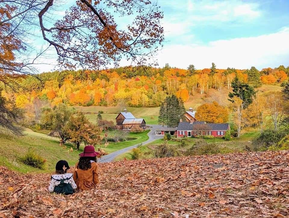 Two children sit in a vast pile of leaves looking over a small farm gilded with fall foliage near Stowe, one of the best places to visit for fall in New England with kids.