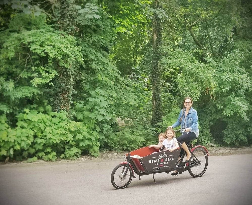 A mother sits atop a bike with a front buggie, which holds her two young girls. All look at the camera with huge smiles.