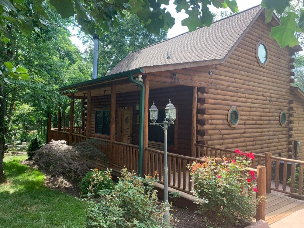 A quaint cabin sits in rural Virginia with beautiful landscaping, as well as lush green trees stationed around the cabin.