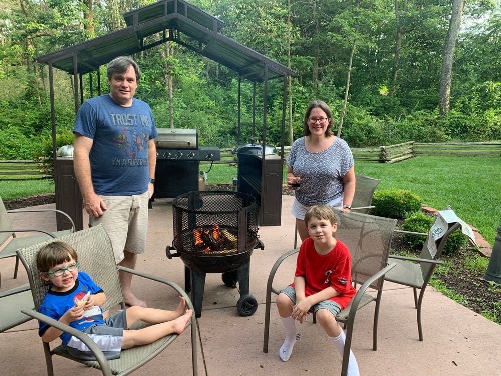 Two young boys sit in lawn chairs with their parents standing behind them, all are smiling. Between the family is a fire pit with a lit fire.