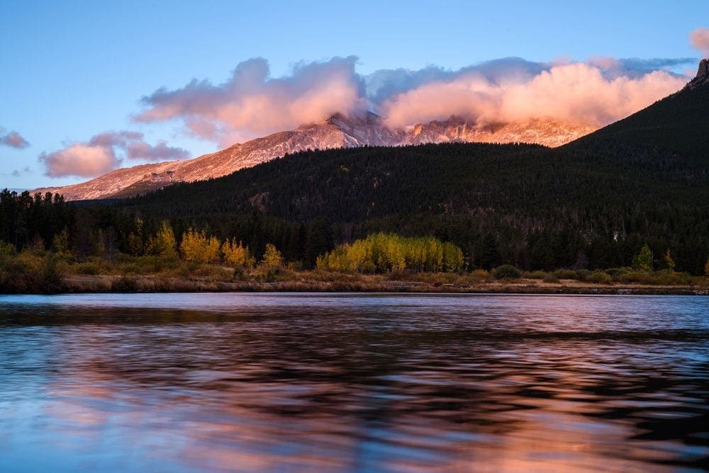 A clear lake reflects a pink sunset, while several mountains stand in the background.