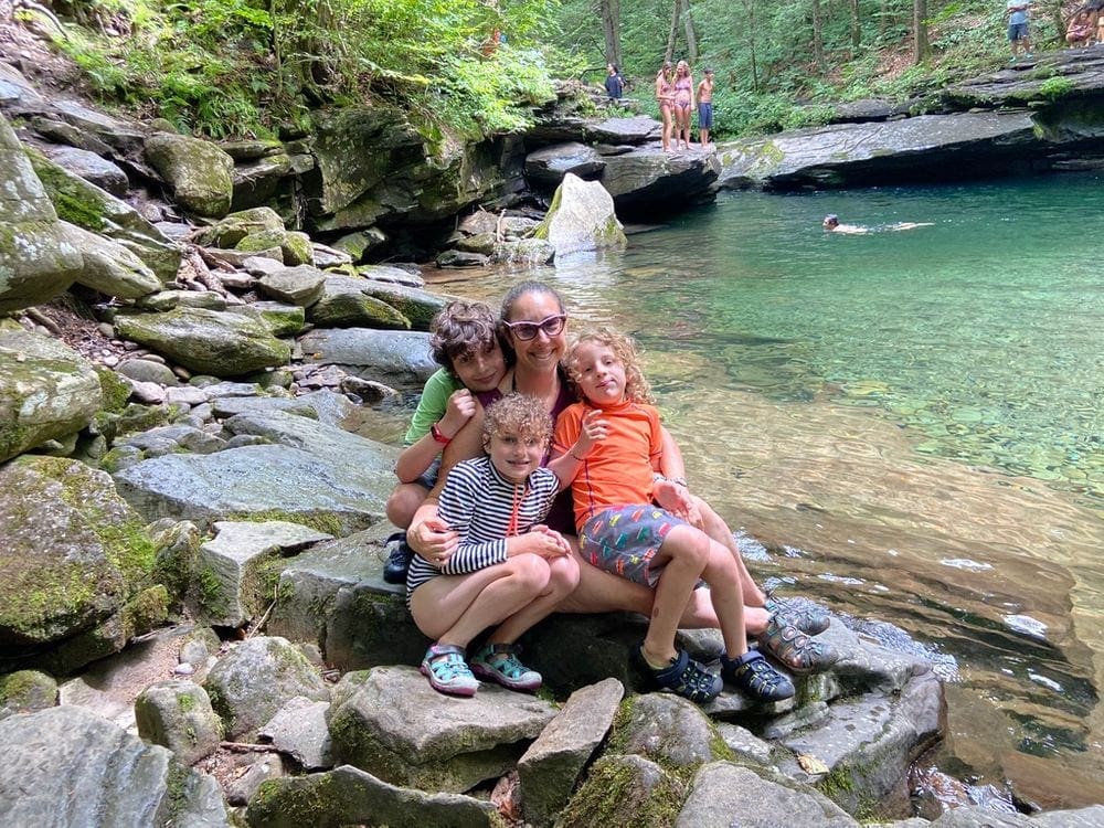 A mother sits on the rocks, closely surrounded by her three children while at Peekamoose Blue Hole. The turquoise waters of the plunge pool is show on the right.