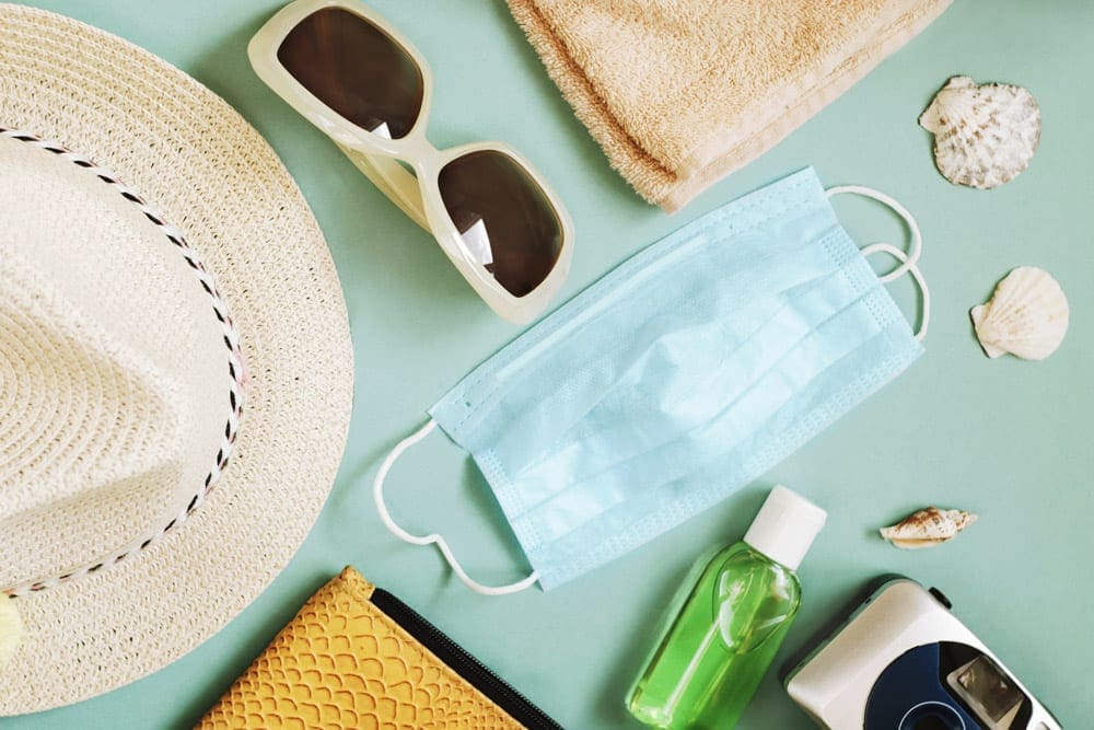 An assortment of items are shown against a green background, including a hat, a mask, sunglasses, and a bottle of hand sanitizer.