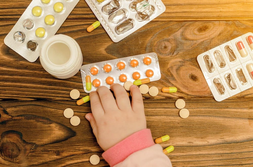 A little hand reachines for packets of medication displayed on a table. Remembering prescription medication is a critical component to any travel first aid kit for kids.