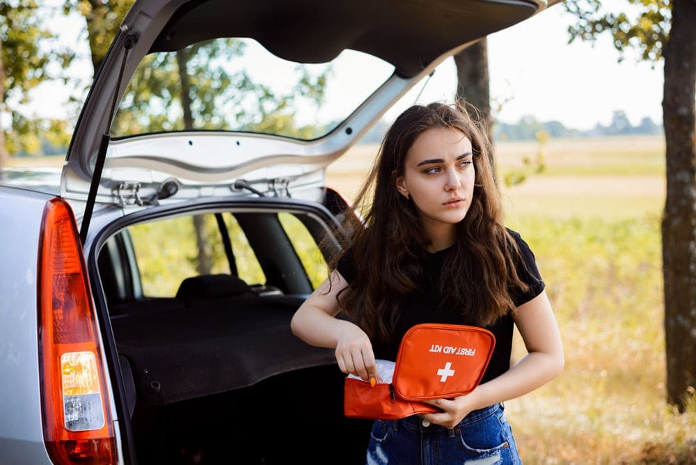 A young woman holding a first aid kit stands behind her vehicle with its hatch open.