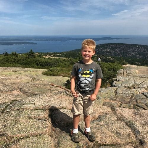 A small boy stands with a stick in his hand with a view of Acadia National Park in the background.
