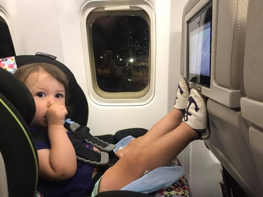An infant girl suck her thumb while sitting in a carseat on a plane preparing to take off.