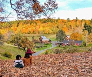 Two children sit in a vast pile of leaves looking over a small farm gilded with fall foliage.