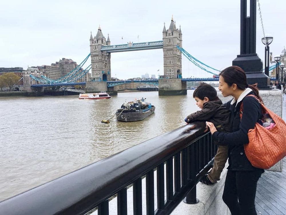 A mother holds her young son agains a guard rail looking over into the Thames River, while the London Bridge stands in the background.