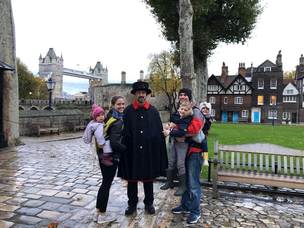 A family of five stands with a guard within the London Tower grounds. Behind them, is several buildings on the grounds, as well as the London Tower in the distance.