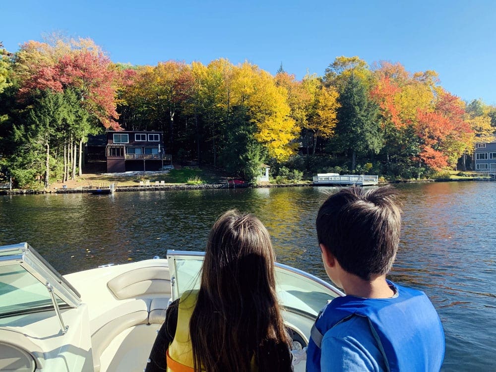 A young boy and girl look toward a stunning fall foliage across a lake from the boat they are riding in.