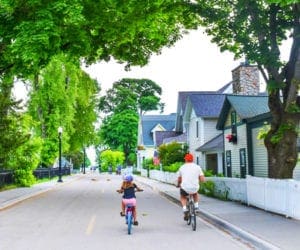 A young girl and her father bike along an enchanting, tree-lined streen on Mackinac Island, Michigan, one of the most charming towns to visit with kids.