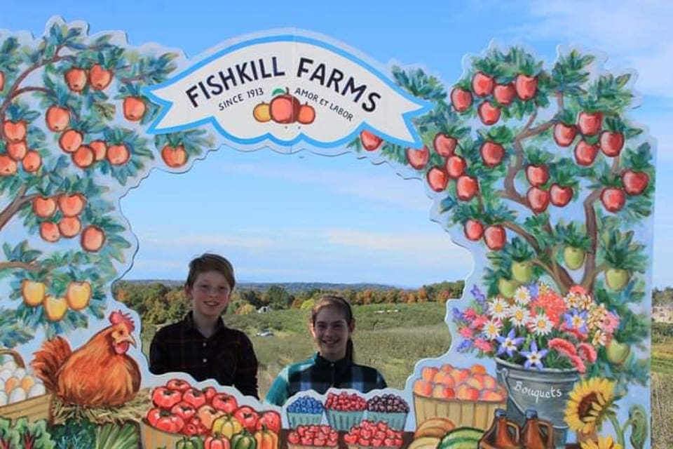 Two kids stand behind a cardboard cutout photograph display featuring apples, pumpkins, and sunflowers at Fishkill Farm.