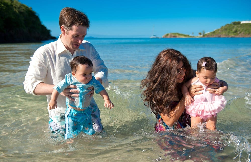 Parents stands in the Caribbean ocean, while dunking the toes of their twin babies in the water.