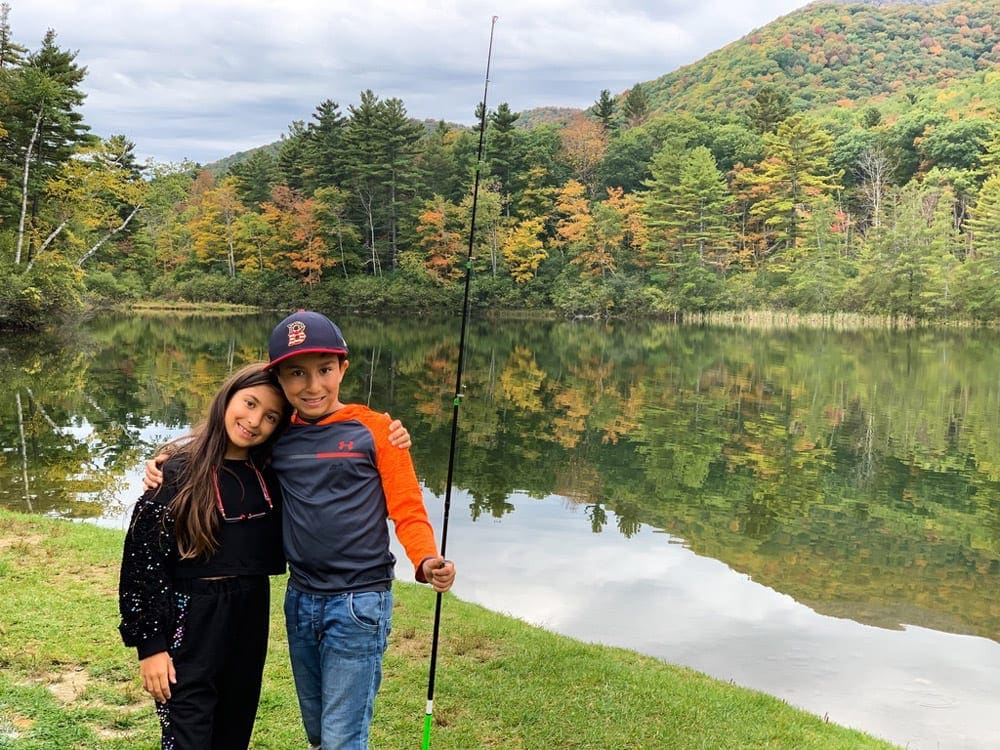 Sister and brother stand close together, while the boy holds a fishing pole. Behind them is a glimmering lake flanked on the far bank with a full array of striking fall colors.