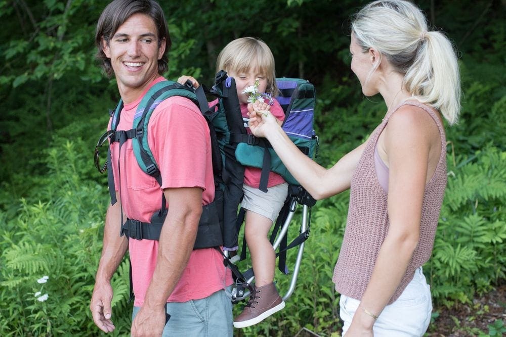 A mother holds up a flower for her child to smell, while he is strapped to the bather of his father on a hike.