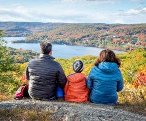 A family of three sits upon a large boulder overlooking a coloful autumn display in New England.