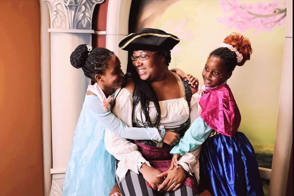 A black mother dressed as a pirated sits smiling at her two daughters, one dressed as Elsa, the other as Anna at Walt Disney World, one of the best places to visit with your young daughter in America.