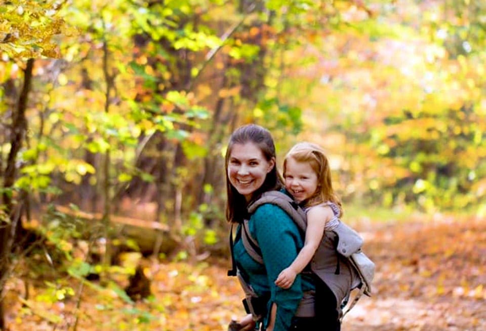 A young girl rides on her mothers back during a brightly colored fall hike in Vermont.