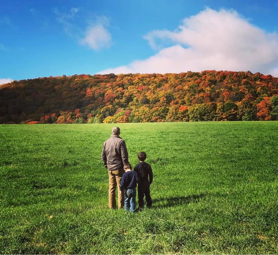 A father stands with his two sons gazing upon a moutain awash in fall hues of orange, yellow, and auburn.