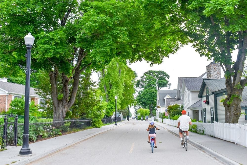 A young girl and her father bike along an enchanting, tree-lined streen on Mackinac Island, Michigan, one of the most charming towns to visit with kids.