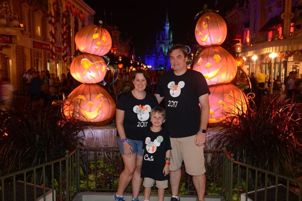 Two parents and their young son stand smiling in front of a jack o'lantern display at Mickey's No So Scary Halloween Party at Walt Disney World.