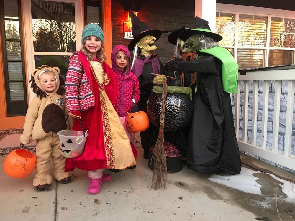 Several kids stand on a porch dressed in Halloween costumes, including a lion, a witch, and several other costumes.
