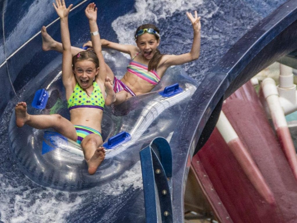 Two girls shoot down a water slide with arms in the air.