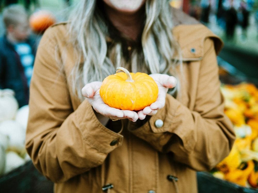 A woman wearing a brown coat holds out a small pumpkin.