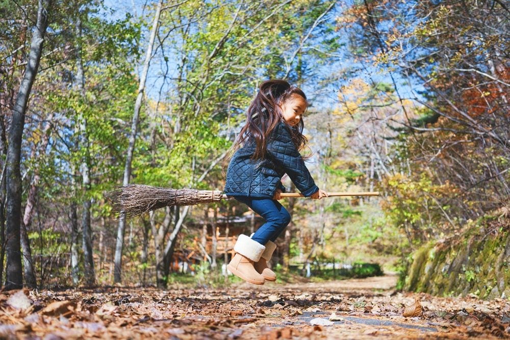 A small Asian girl jumps in the air, pretending that she is flying on a broom during a crisp, fall day.