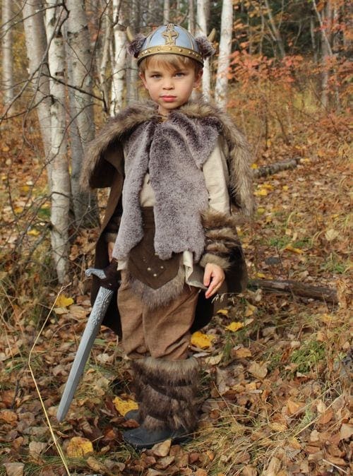 A young boy wear fur pelts and a viking hat poses in the woods, one of the best travel themed costumes for kids.