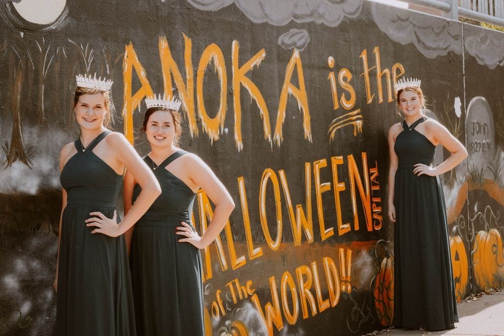 Three women wearing black dresses and crowns stand by a graffiti sign reading "Anoka is the Halloween Capital of the world!!".