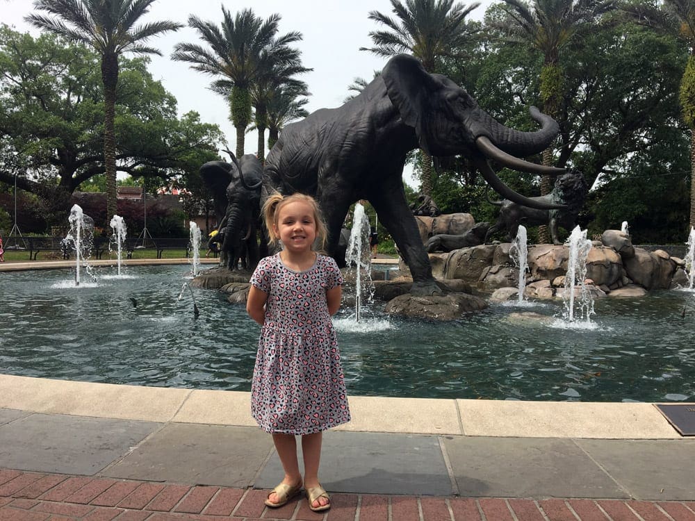 A young girl stands smiling in front of a fountain of elephants at the Audubon Zoo.